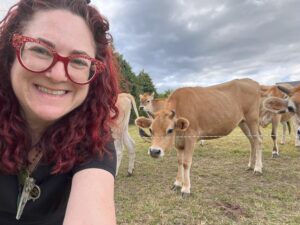 Person with long curly hair and red glasses taking a selfie of herself with brown cows behind her.