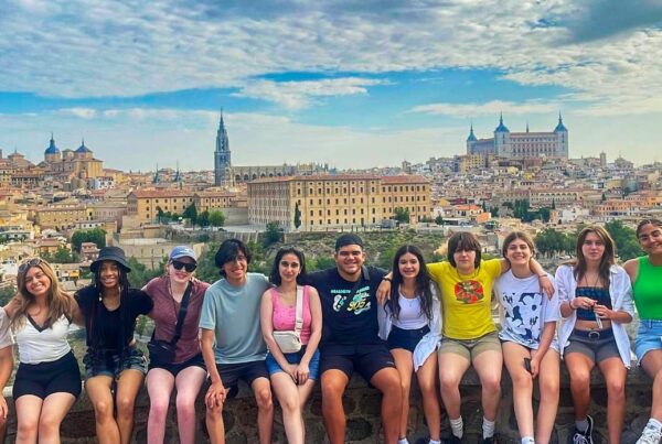 Thirteen students sit on a stone ledge with their arms around each other. In the background is a large city with historic architecture. The sky is bright blue with large white clouds.