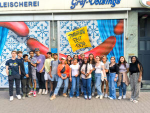 Large group of high school students standing in front of a colorful mural of giant sausages.