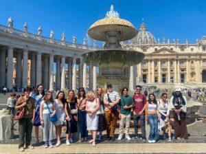 Group of 14 high school aged students stand in front of a large marble fountain. Behind them is a large plaza with the dome of St. Peter's Basilica in the distance.