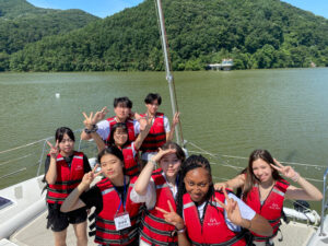Eight high school students are on a boat on a lake. They are all wearing red life vests and giving the peace sign. Off in the distance are tree covered hills.