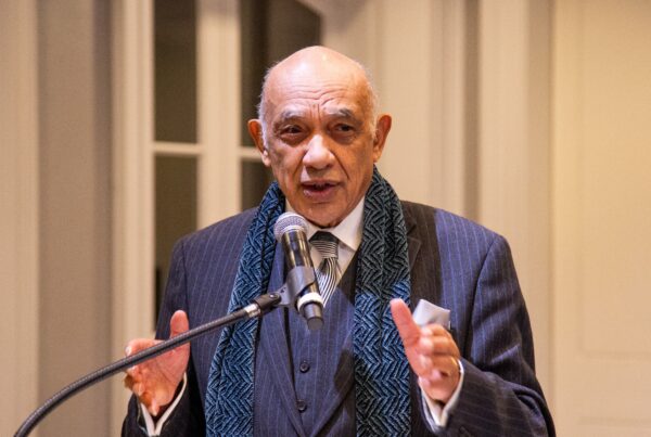 Male standing at a podium talking into a microphone. His hands are raised, gesturing.