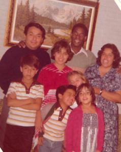 An Experiment host family in Mexico poses with an Experiment students. There are four young children and two parents. They are wearing clothing from the 1970s, and there is a framed picture of a river landscape behind them. 