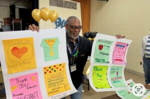 A man is holding up two posters with colorful drawings taped to them. Around his neck is a school badge and behind him are balloons.