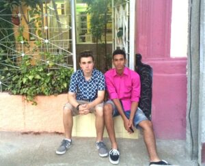 Two male Experiment high school students sit on a stone wall. The wall is yellow and the wall behind them is pink.